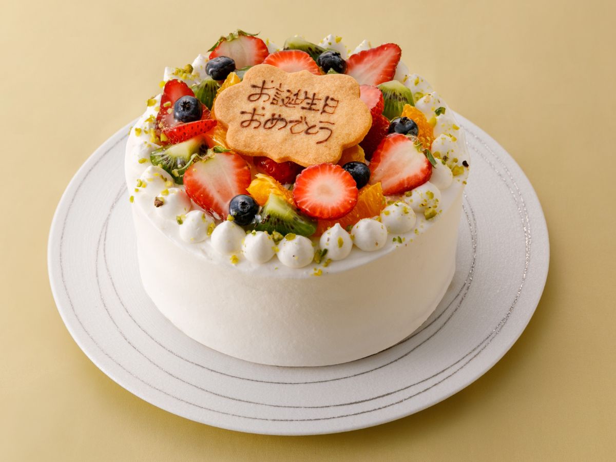 写真：季節のフルーツがトッピングされた華やかなケーキ。ケーキの上には「お誕生日おめでとう」と書かれたクッキーが乗っている。